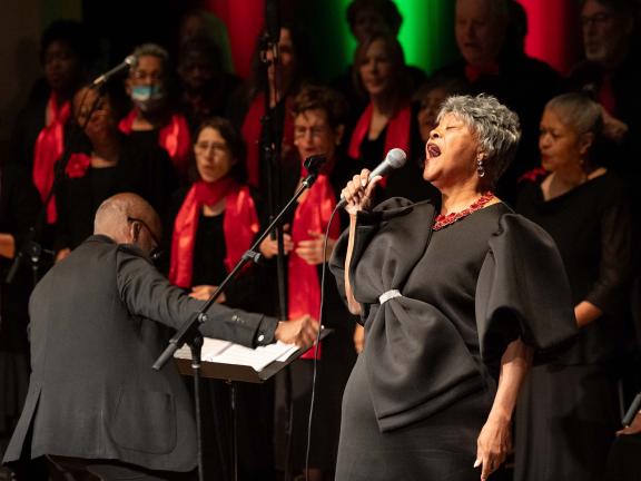 A woman holds a microphone to her mouth and sings while behind her a man conducts a choir.