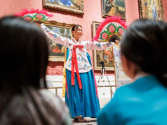 A woman in traditional dress holds fans and dances in front of an audience.