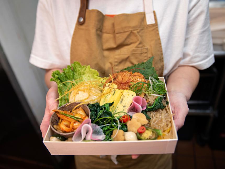 A chef holds up a box full of various foods—lettuce, kimchi, egg, noodles, and seaweed