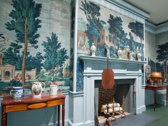 An interior with ornate wallpaper showing trees and Roman architecture. The room is filled with tables, on top of which sit decorative ceramics, and a fireplace has birch logs in it.