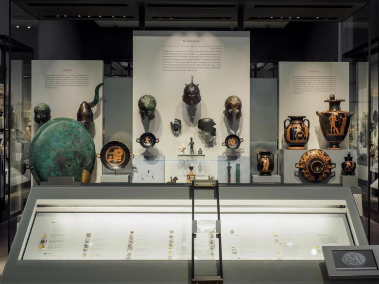 Display case filled with ancient Greek coins in foreground, with helmets and other military depictions in display case in background