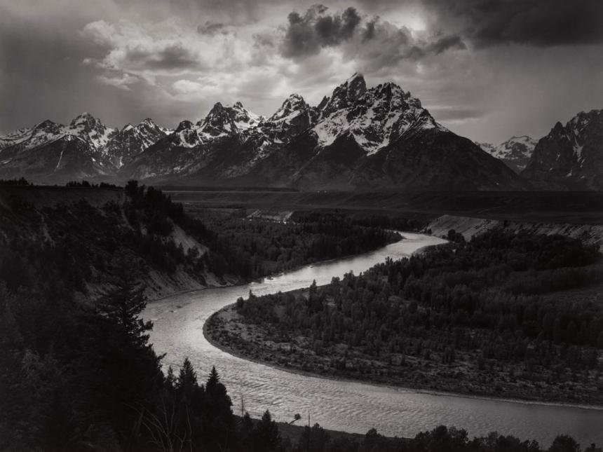 black and white landscape photograph depicting river snaking through forest with huge mountains in the background