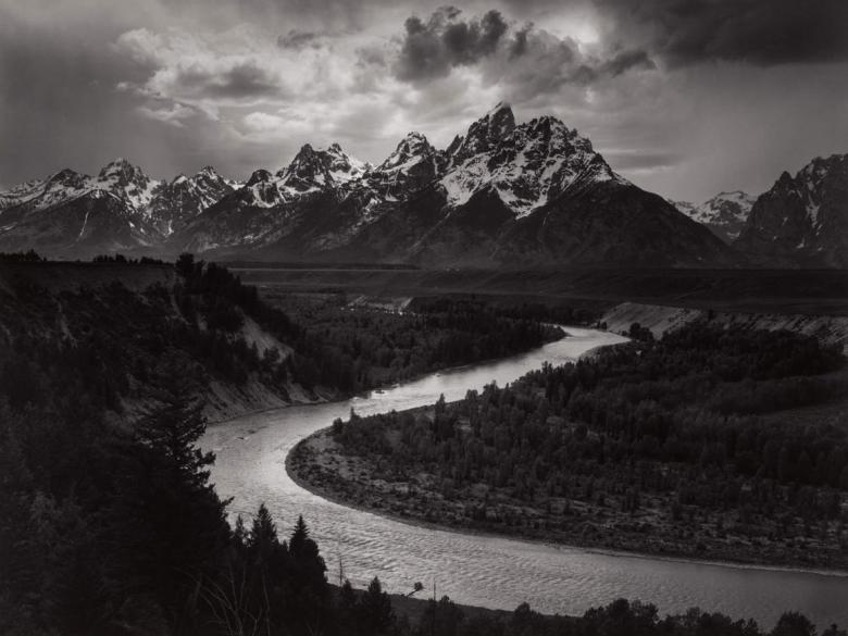 black and white landscape photograph depicting river snaking through forest with huge mountains in the background