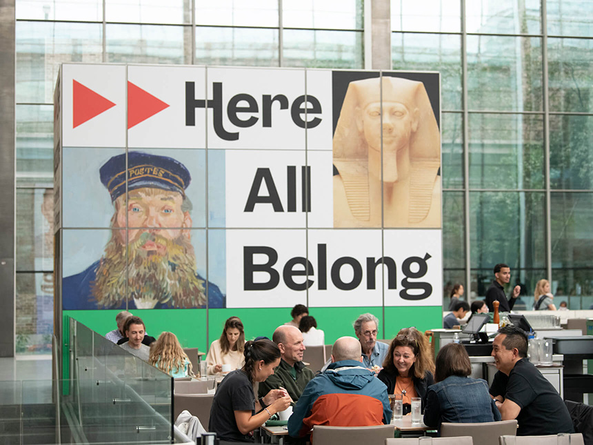 Diners sitting at tables in New American Cafe in Shapiro Family Courtyard, with large sign "Here All Belong" in the background