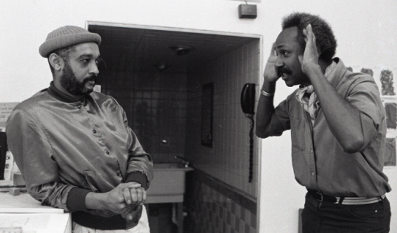 Two people stand across from each other; one holds his hands up to his face, the other leans against a ledge.