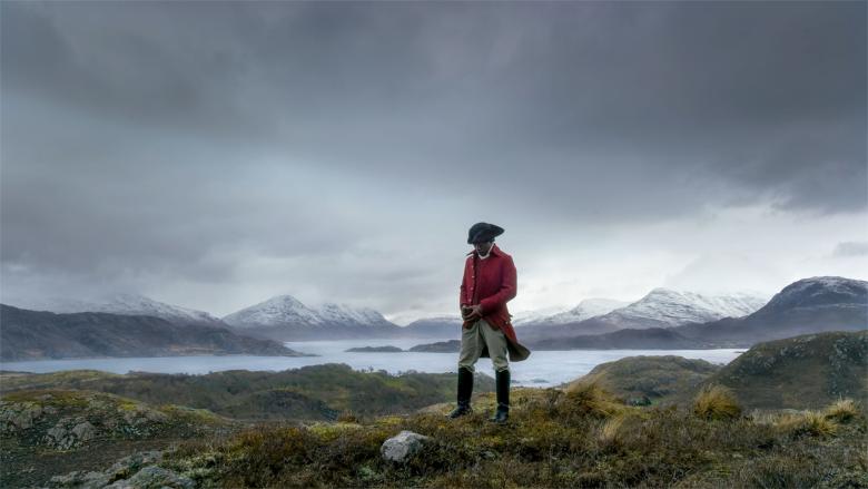A man in a red coat stands on a beach beneath a dramatic cloudy sky.