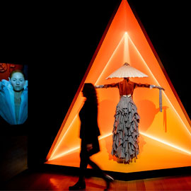 Visitor walking by orange-lit display of outfit in Gender Bending Fashion exhibition