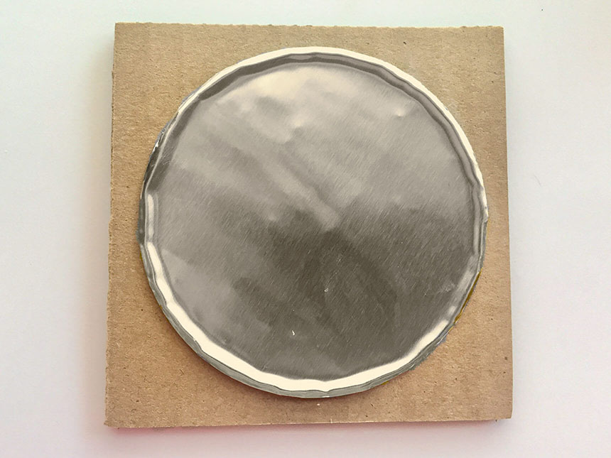 circular piece of foil placed placed on top of square piece of cardboard, with the foil's gold side facing down