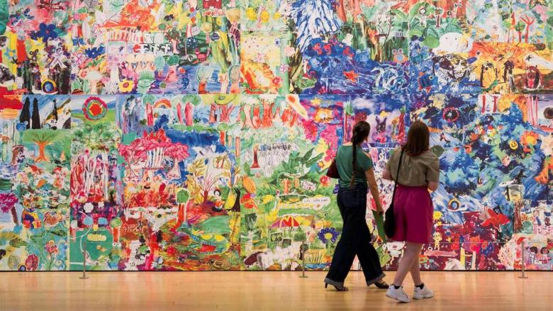 Two visitors look at large gallery wall covered with colorful paintings from kids made for Community Arts Initiative's 2025 Artist Project, Into the Forest