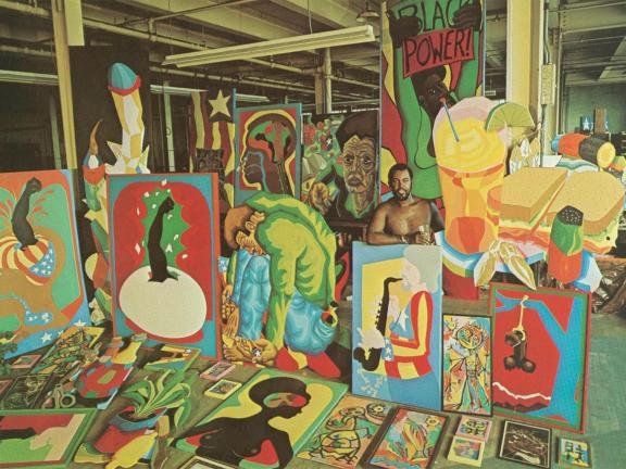 A shirtless man stands in a warehouse-like space surrounded by colorful paintings.