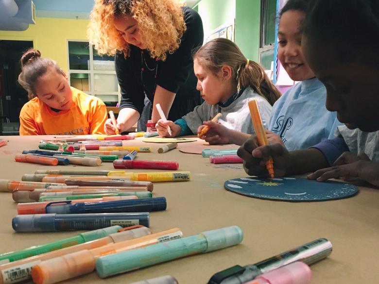 Kids sitting around table working on art project with markers, guided by teacher