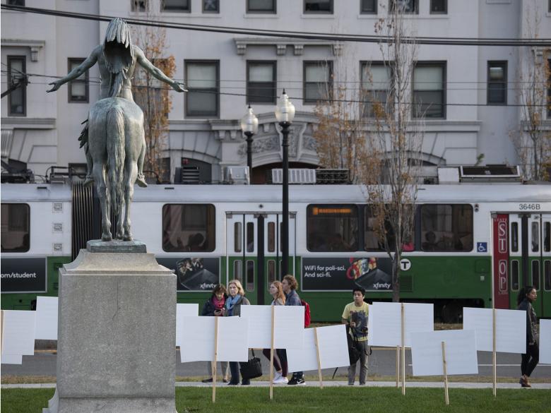 A sculpture of a Native American man on horseback stands on a pedestal in front of a passing MBTA Green Line trolley. Signs surround the sculpture with visitor thoughts on the work of art.
