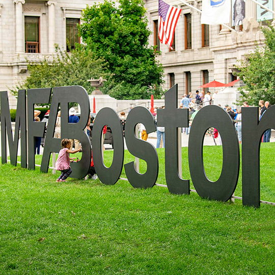 Wooden letters spelling out MFA Boston on Huntington Avenue Entrance lawn, with small child playing in between letters