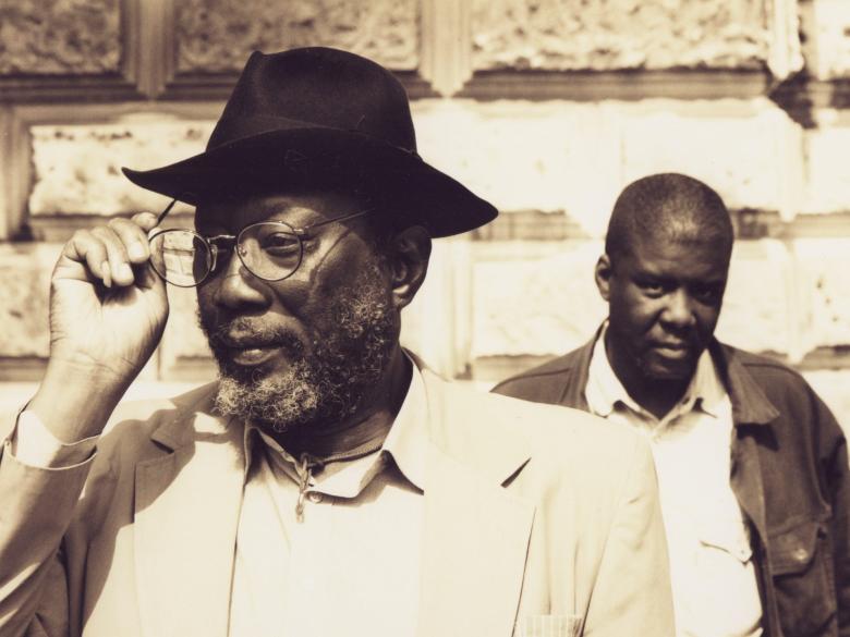 Frank Bowling stands in the foreground and adjusts his glasses as Eddie Chambers stands behind him. 