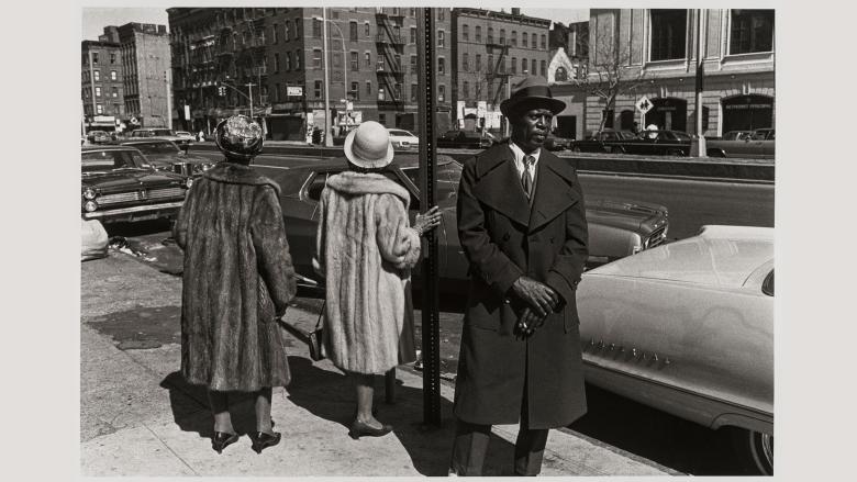A man in a suit and had stands looking in the distance; behind him, two women in fur coats turn their backs.