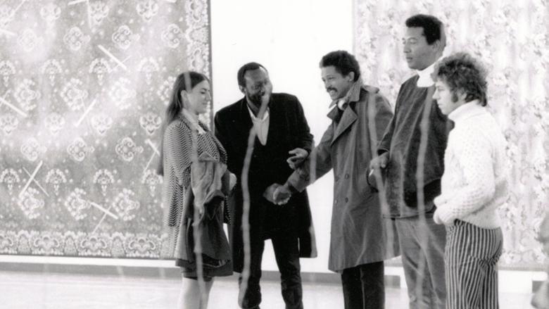Four people stand in a line against a white wall with large tapestries against it.  Black-and-white image.