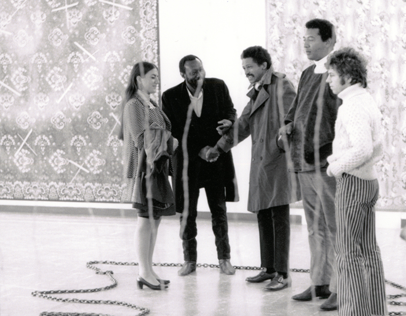 Four people stand in a line against a white wall with large tapestries against it.  Black-and-white image.
