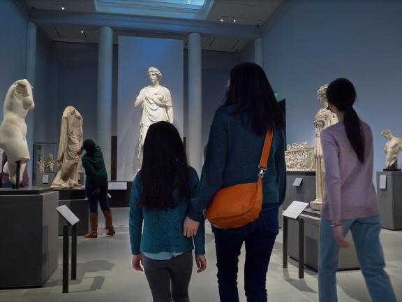 Parent with two children walking through large gallery with sculptures of various Greek gods and goddesses on pedestals