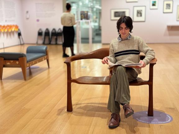A visitor reads an object guide on a wooden bench in "Tender Loving Care"