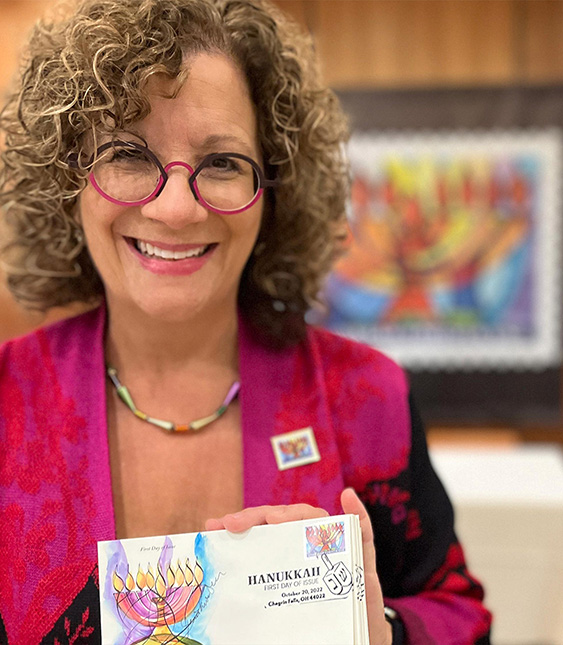 Artist Jeanette Kuvin Oren holding up first day issue of USPS's 2022 Hanukkah stamp