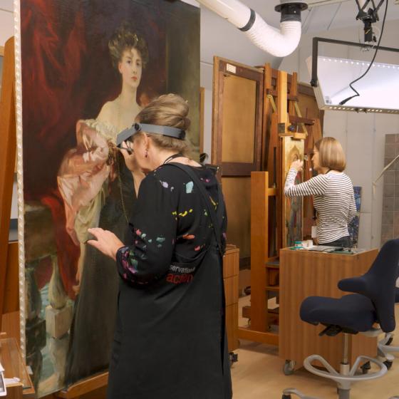 Conservator standing in front of painting on easel; the painting depicts woman in long black dress and pink, billowy wrap