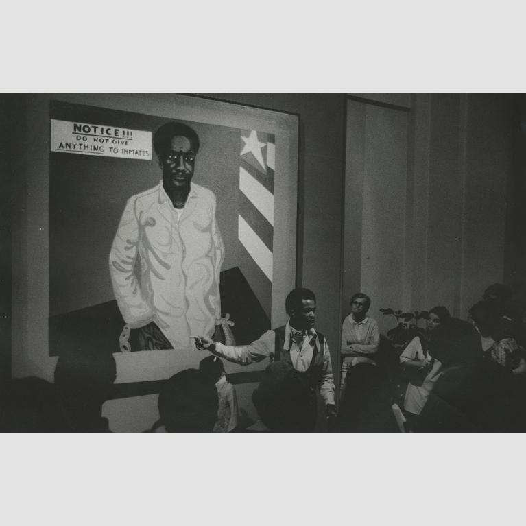 A man stands in front of a large painting of a many and speaks about it to a rapt audience.