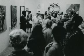 A crowd of people surround a man gesticulating with his arms as he stands and speaks in front of a wall of paintings.