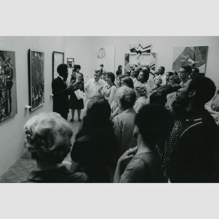 A crowd of people surround a man gesticulating with his arms as he stands and speaks in front of a wall of paintings.
