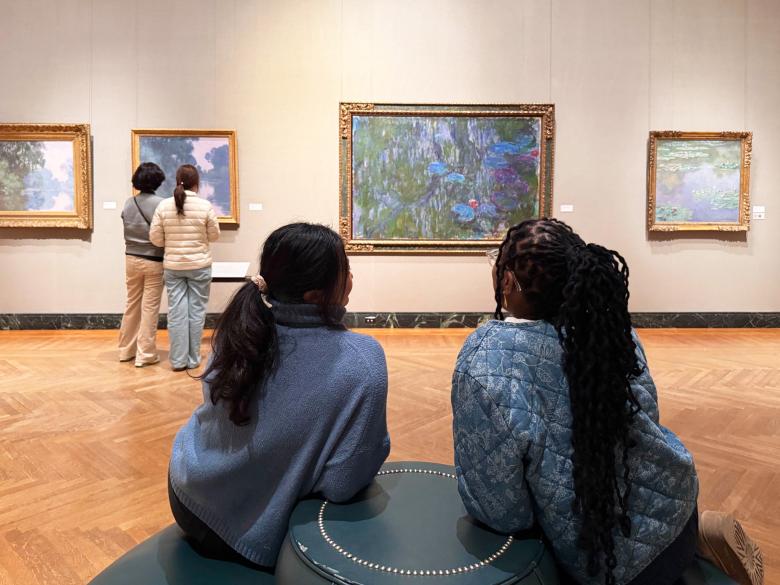 Two people sit on a bench and face a large colorful painting framed and hung on a wall among other paintings.
