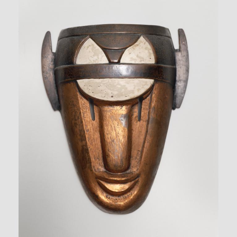 A copper and enamel mask mimicking a human face wears a white and dark brown headpiece.