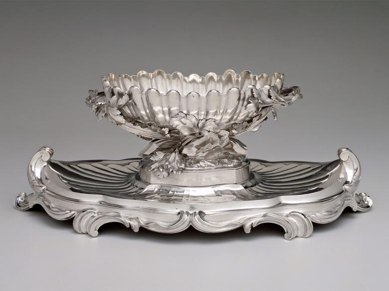 An ornate silver suaceboat sits atop a lavishly decorated stand.