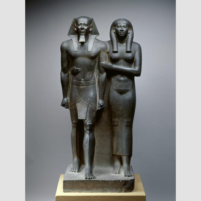 Dark grey statue of an Egyptian king and his wife with her arms wrapped around her husband.