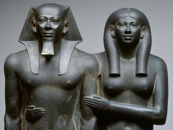 Dark grey statue of an Egyptian king and his wife with her arms wrapped around her husband.