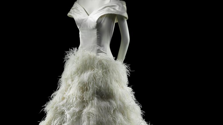 A gray mannequin wears a shiny white dress with ruffled plumes from the hip down.
