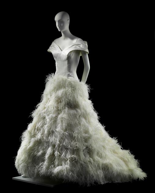 A gray mannequin wears a shiny white dress with ruffled plumes from the hip down.