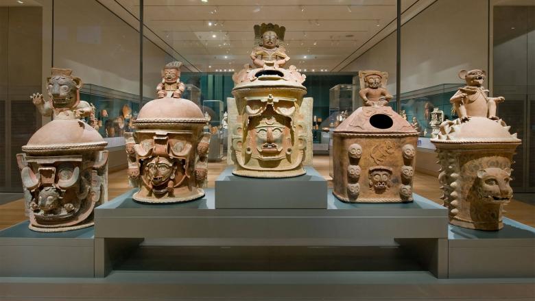 View of six Mayan urns arranged in a row inside a gallery case