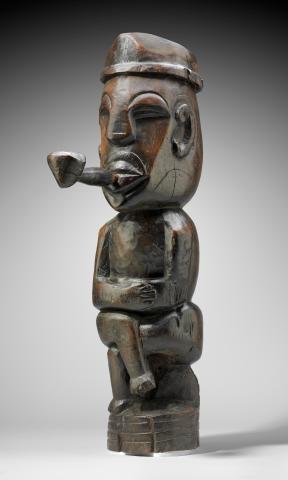 A wood sculpture of a man sitting cross-legged on a stump, wearing a hat and smoking a pipe. 