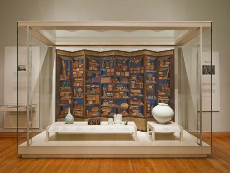 View of display case in Arts of Korea gallery, featuring pottery on low tables in front of screen depicting bookcase