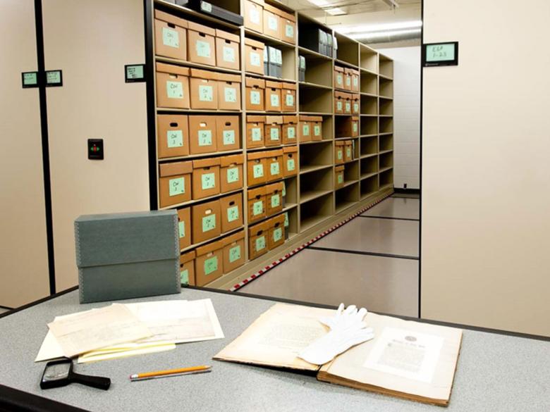 Library stacks with archival boxes, a table with old documents spread out across it.