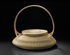 A squat basket with a handle, woven from different types of plants. 