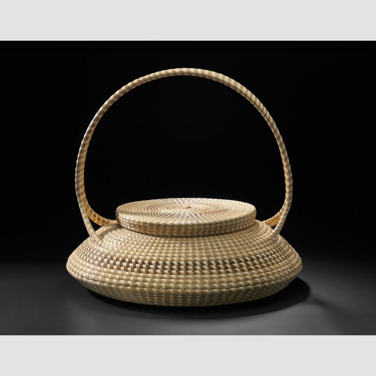 A squat basket with a handle, woven from different types of plants. 