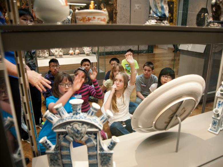 School children sit on the ground and raise their hands as they view artwork in a glass case. 