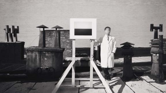 A black-and-white photograph of a man in a white lab coat with a blurred face standing beside an easel on a rooftop.