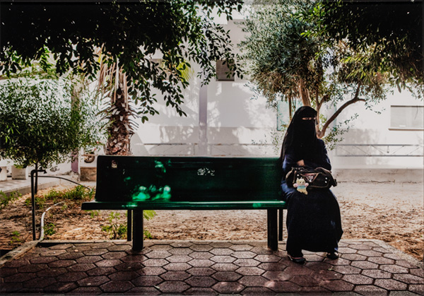 Wearing a full face covering, a woman sits on a shaded bench in a sunny courtyard and holds a backpack.