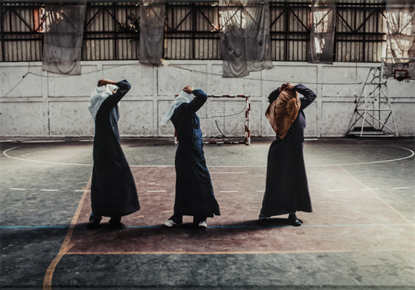 Three women with head coverings walk in a gym with their arms raised above their shoulders and hand resting on the top of their heads.
