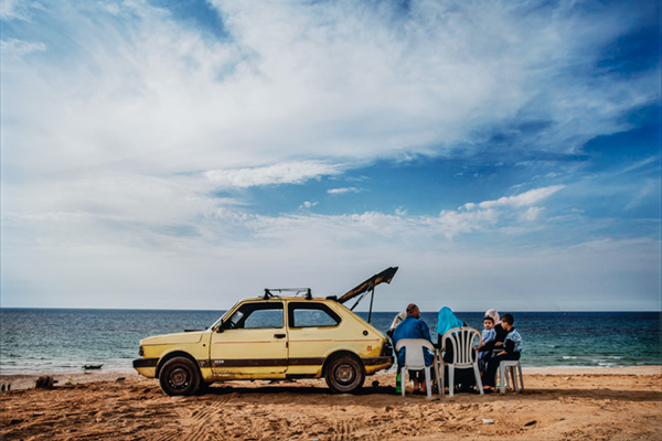 A group of people sit in plastic chairs behind a beat-up yellow car on a beach with a bright, overcast sky above and the ocean in the distance.