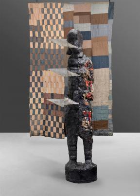 Quilt with tan, blue and brown squares and rectangles with a sculpted figure in front of it.