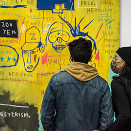 Two visitors wearing masks standing side-by-side and looking at the brightly colored Basquiat mural