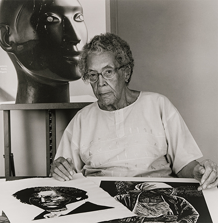 An elderly woman sits at a table with artwork in front of and behind her.