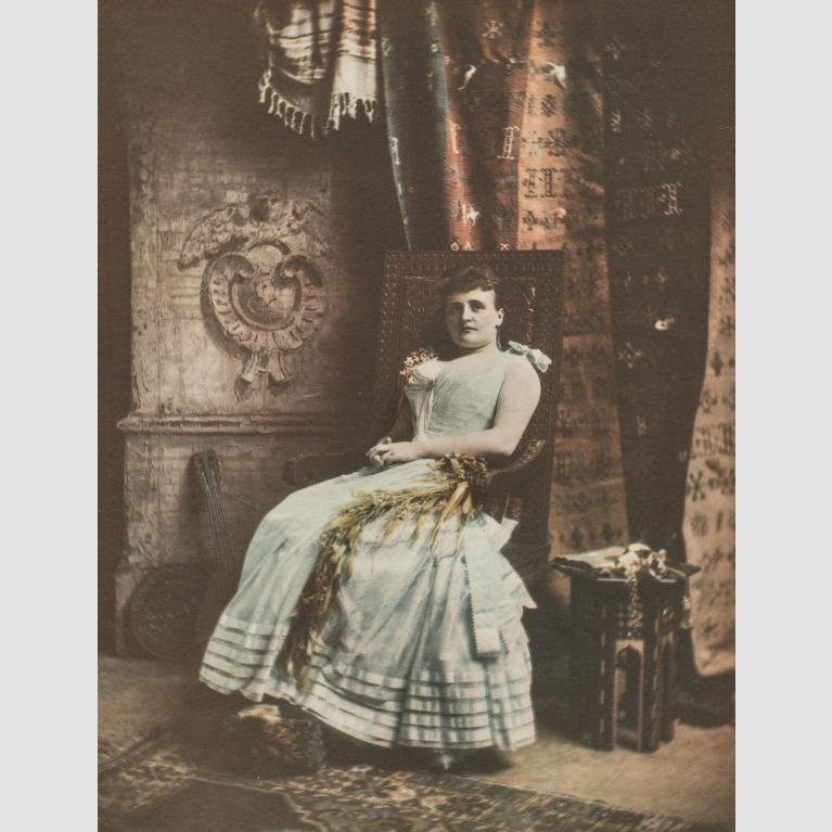 A portrait of a woman in a gown, sitting in a chair against a backdrop of curtains and blankets.